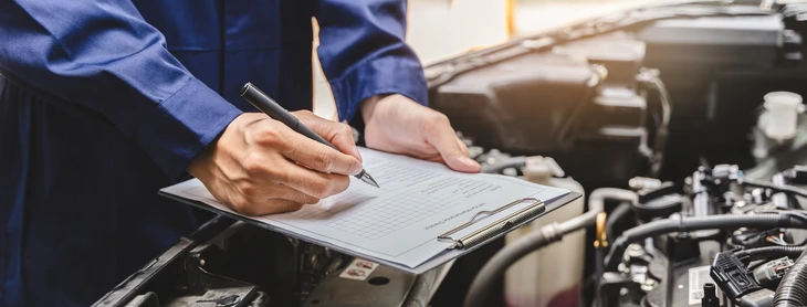 Technicien en combinaison grise et masque FFP2, agenouillé à l'arrière d'une Peugeot 3008 grise dans un garage de contrôle technique