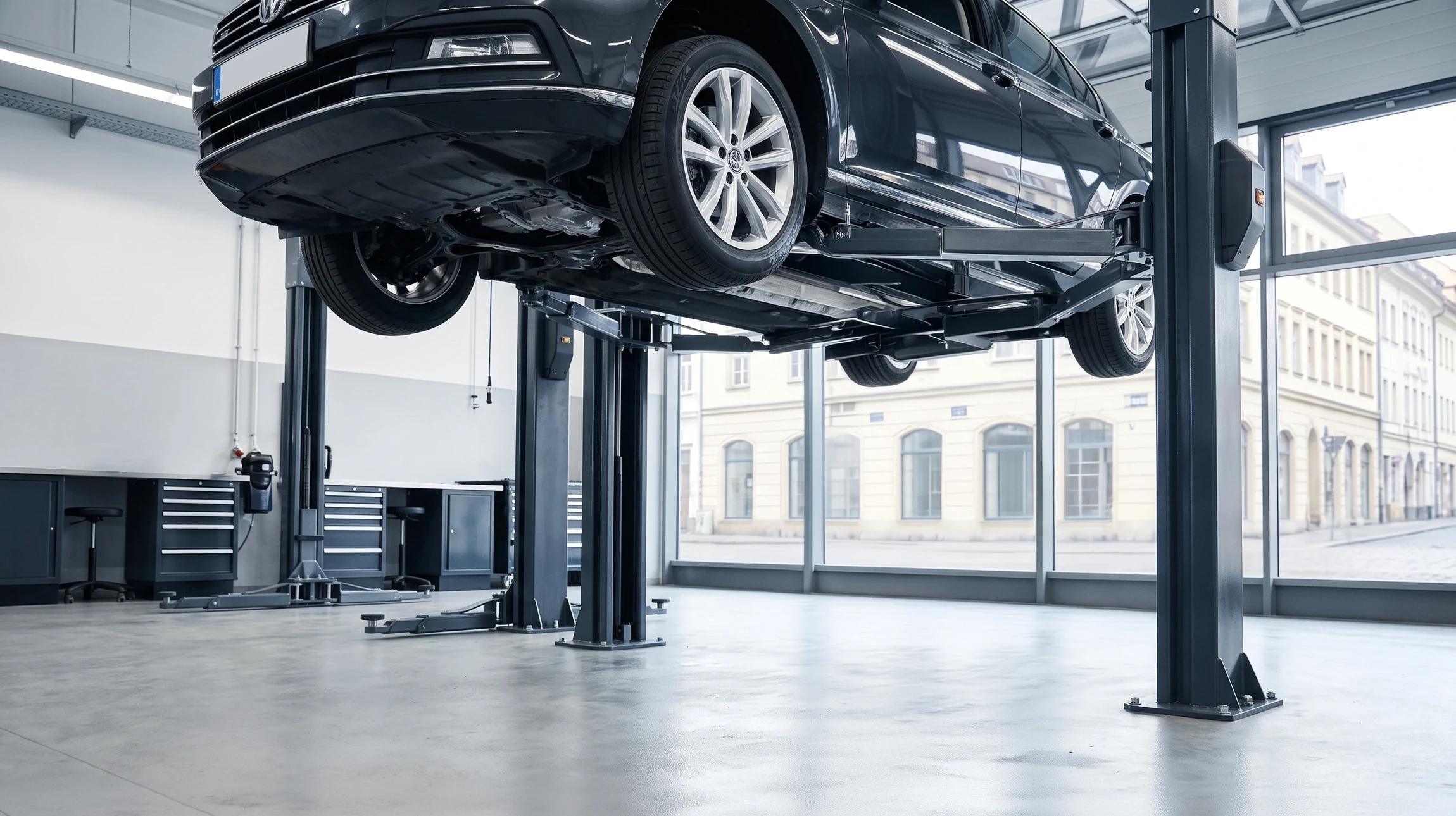 Technicien de contrôle technique automobile en combinaison de travail grise dans un atelier professionnel, effectuant une vérification sous un véhicule surélevé sur pont élévateur. Le technicien connecte des équipements de diagnostic ou effectue une inspection visuelle des éléments de sécurité (freins, suspension, direction). L'environnement montre un atelier moderne et équipé avec équipements de diagnostic professionnels. La scène transmet la confiance, l'expertise technique et la rigueur du contrôle automobile.