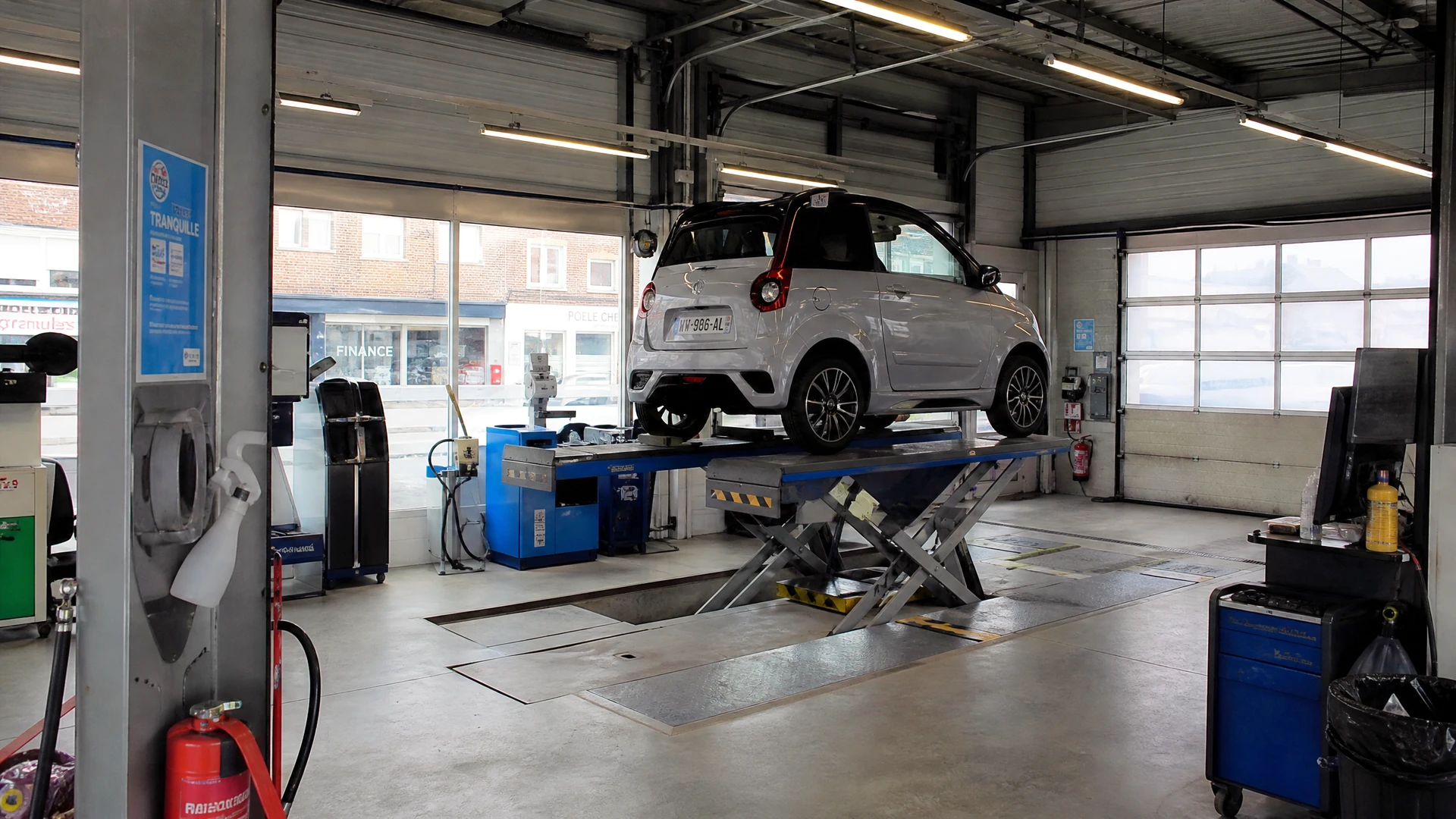 Technicien en combinaison grise et masque FFP2, agenouillé à l'arrière d'une Peugeot 3008 grise dans un garage de contrôle technique